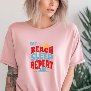 May include: A pink t-shirt featuring a graphic design that reads "Eat, Beach, Sleep, Repeat" in a retro style with a sun and waves.