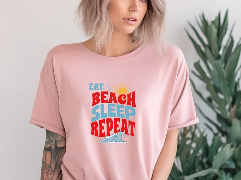 May include: A pink t-shirt featuring a graphic design that reads "Eat, Beach, Sleep, Repeat" in a retro style with a sun and waves.