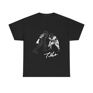May include: A black t-shirt featuring a graphic illustration of two football players wearing jerseys with the number 12. One player wears a black uniform and the other wears a white uniform. Below the players is a white signature that reads T. Hunter.