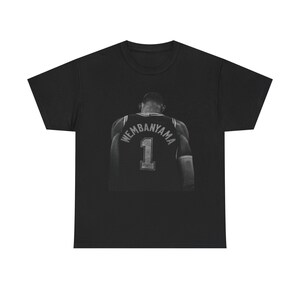 May include: Black t-shirt featuring a grayscale image of a basketball jersey with the name "Wembanyama" and the number "1" on the back. The jersey is set against a dark background, creating a striking contrast.