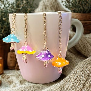 May include: A pink mug with four colorful mushroom tea infusers hanging from the rim. The mushroom caps are blue, pink, purple, and orange, each with a different pattern. The infusers are suspended by gold chains.