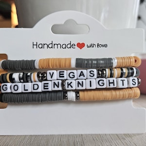 May include: Stack of four handmade bracelets on a white display card. The bracelets are made of gray, tan, and white disc beads. One bracelet spells out "VEGAS" and another spells out "GOLDENKNIGHTS" with white letter beads.