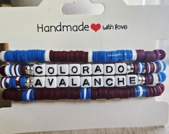 May include: Handmade bracelets featuring blue, white, and maroon disc beads. The bracelets spell out "COLORADO AVALANCHE" with white letter beads. The card reads "Handmade with love" with a red heart.
