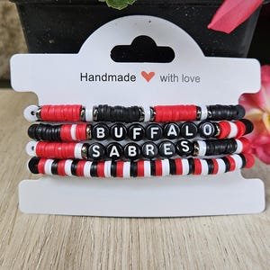 May include: Stack of four beaded bracelets in red, black, and white. One bracelet spells out "BUFFALO SABRES" with black letter beads. The bracelets are displayed on a white card with the text "Handmade with love".