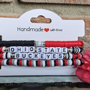 May include: Stack of beaded bracelets in black, red, white, and gray. One bracelet spells out "OHIO STATE BUCKEYES" with white letter beads. The bracelets are displayed on a white card that reads "Handmade with love".