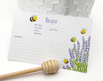 May include: A recipe card with a lavender and bee design. The card has lines for ingredients, dish name, prep time, cook time, serves, and references. The word "Recipe" is at the top. A wooden honey dipper is in the foreground.