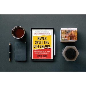 Könnte beinhalten: Eine flache Ansicht eines Schreibtisches mit einem Buch mit dem Titel "Never Split the Difference: Negotiating as if Your Life Depended on It" von Chris Voss, einer Tasse Kaffee, einem Stift, einem Notizbuch, einer kleinen Schachtel Tee und einem Metallsieb.