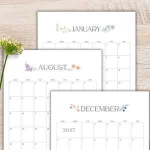 May include: Three white calendar pages for the year 2027, featuring the months of January, August, and December. Each page displays a grid with days of the week and delicate watercolor floral illustrations in soft pastel colors flanking the month names.