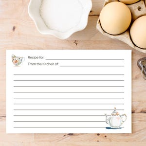 Puede incluir: Una tarjeta de receta blanca con espacios rayados, con ilustraciones de acuarela de una taza de té y una tetera. La tarjeta incluye indicaciones para la receta y la cocina de donde proviene. Huevos, un cuenco de azúcar y cucharas medidoras están en el fondo.