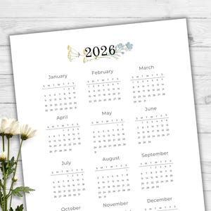May include: A white 2026 calendar with floral accents. The calendar displays months from January to December, with each month's days arranged in a grid. The year "2026" is at the top, with decorative flowers. The calendar is on a white wooden surface.