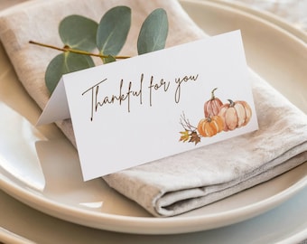 May include: A white place card with the text "Thankful for you" in brown script, featuring watercolor pumpkins and fall foliage. The card rests on a linen napkin and a white plate, with eucalyptus sprigs.