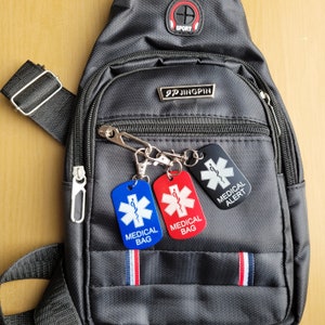 May include: A black crossbody bag with a zipped pocket and a silver buckle. The bag has three medical alert tags attached to it. The tags are red, blue and black with white text. The red tag says "MEDICAL BAG", the blue tag says "MEDICAL BAG", and the black tag says "MEDICAL ALERT".