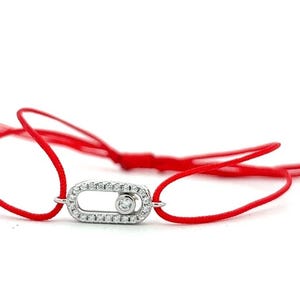 May include: A red cord bracelet featuring a silver-toned oval-shaped charm encrusted with small clear stones. The charm contains a single, central, circular clear stone that is set within a movable inner track, creating a dynamic, sliding effect.