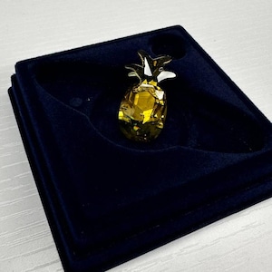 May include: A decorative pineapple figurine with a faceted, yellow crystal body and gold-colored leaves. The pineapple is displayed in a dark blue velvet box, creating a striking contrast. This item is a decorative piece.