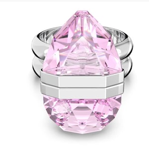 Könnte beinhalten: Ein rosafarbener Kristallring mit einem facettierten, tropfenförmigen Mittelstück. Der Ring hat ein silbernes Band und ein geometrisches Design. Der Kristall ist hellrosa und der Ring hat einen modernen, eleganten Stil.