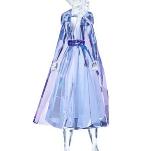 Puede incluir: Una figura de cristal de Elsa de Frozen II de Disney. La figura está hecha de cristal transparente con detalles azules y morados. Elsa lleva su característico vestido y capa azul hielo. Tiene el pelo largo y rubio y sostiene las manos delante de ella.