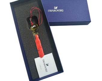 May include: A Swarovski crystal ornament with a red tassel and a gold-colored crystal figure, presented in a navy blue box. The box is open, revealing the ornament and a white tag with the Swarovski logo.