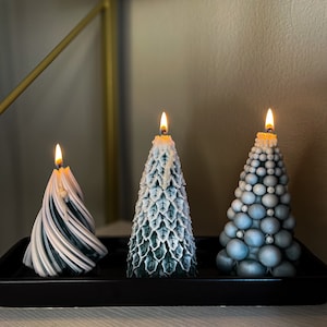 Puede incluir: Tres velas de árbol de Navidad azul verdoso y blanco en una bandeja negra. Las velas están encendidas y tienen diferentes diseños.