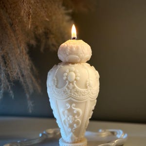 Puede incluir: Una vela blanca, intrincadamente detallada, con forma de jarrón con una llama que arde en la parte superior. La vela está sentada en una bandeja blanca.