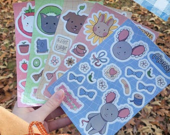 May include: A hand holds a stack of colorful sticker sheets featuring cute animal illustrations. The stickers include a cat, dog, bunny, and mouse, along with various decorative elements. The background shows fall leaves and a decorative star.