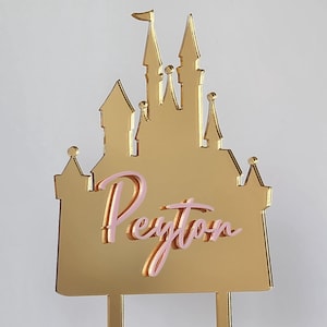May include: A gold-colored castle cake topper with the name "Peyton" in pink script. The castle features multiple turrets and a flag. The topper is designed to be inserted into a cake.