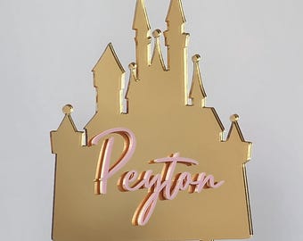 May include: A gold-colored castle cake topper with the name "Peyton" in pink script. The castle features multiple turrets and a flag. The topper is designed to be inserted into a cake.