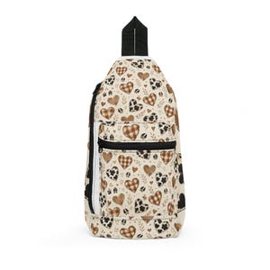 May include: A cream-colored sling backpack featuring a repeating pattern of brown plaid hearts, black and white cow print hearts, and small leaf sprigs. The bag includes a black zipper closure on the front pocket and a black top carry handle.