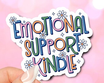 Puede incluir: Una pegatina con el texto "Emotional Support Kindle" en letras de colores con un diseño de margarita. La pegatina está sostenida por una mano con esmalte de uñas blanco.