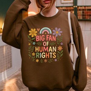 May include: A brown sweatshirt with the text "BIG FAN OF HUMAN RIGHTS" in colorful letters, surrounded by flowers and a rainbow. The sweatshirt is a solid brown color.
