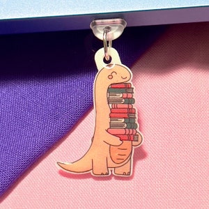 May include: A pink dinosaur bookmark with a stack of colorful books in its mouth. The bookmark is made of acrylic and has a clear plastic loop for hanging.