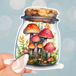 Op de afbeelding: Een glazen pot gevuld met verschillende rode en oranje paddenstoelen, groene planten en gele bloemen. De pot heeft een kurken deksel en staat op een witte achtergrond.