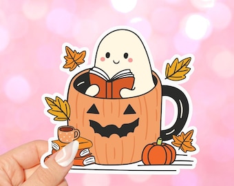 May include: A white ghost reading a book inside a pumpkin-shaped mug with a black handle. The mug is surrounded by fall leaves and a stack of books. There is a small cup of coffee and a small orange pumpkin next to the mug.