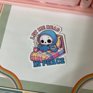Puede incluir: Una pegatina con un esqueleto de dibujos animados que lleva una sudadera azul y lee un libro en la cama. El texto de la pegatina dice "D&eacute;jame leer en paz."