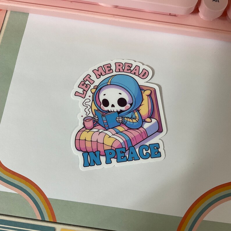 Puede incluir: Una pegatina con un esqueleto de dibujos animados que lleva una sudadera azul y lee un libro en la cama. El texto de la pegatina dice "D&eacute;jame leer en paz."