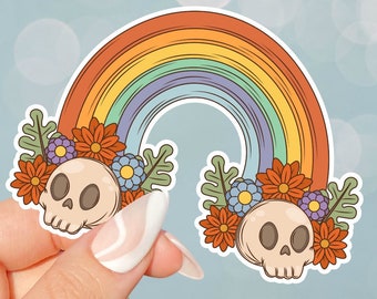 May include: A sticker featuring a rainbow arch with two cartoon skulls and floral accents. The rainbow is in shades of orange, yellow, green, blue, and purple. The flowers are orange, red, purple, and blue.