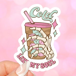 May include: A sticker with a cartoon illustration of a skeleton hand holding a cup of cola. The text "Cold like my soul" is written in pink letters on the sticker.