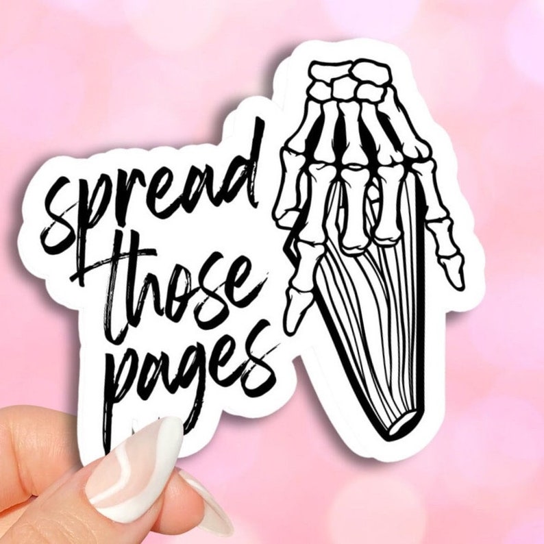 The image shows a sticker with the text "Spread Those Pages" written in black. A black and white drawing of a skeleton hand holding a book is next to the text. The sticker is being held by a hand with painted nails.