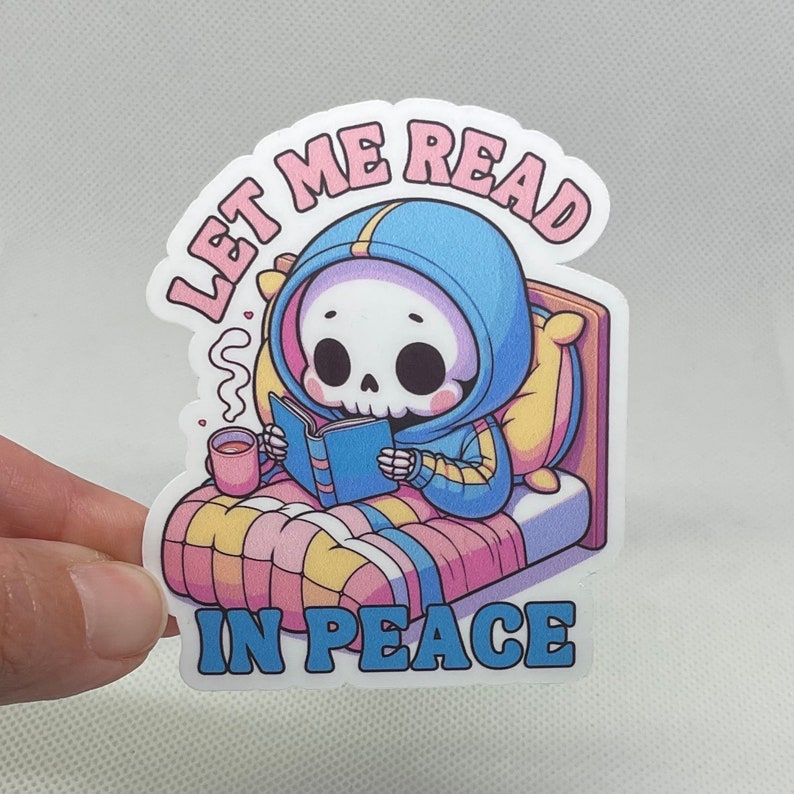 Puede incluir: Una pegatina con un esqueleto de dibujos animados que lleva una sudadera con capucha azul y lee un libro en la cama. La pegatina dice "D&eacute;jame leer en paz".