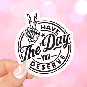 May include: Black and white sticker featuring a skeleton hand making a peace sign. The sticker reads "Have the Day You Deserve".