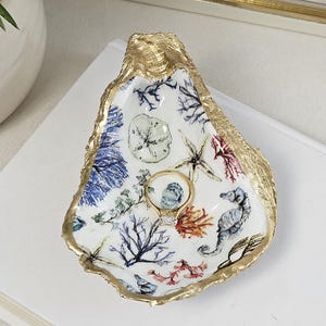 May include: A decorative oyster shell ring dish with a gold-painted edge. The interior features a white background with a colorful sea life design, including coral, seahorses, and starfish. A gold ring with a diamond sits in the center.