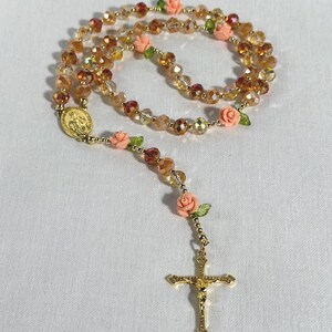 May include: A rosary featuring amber and clear beads, a gold-tone crucifix, and peach-colored rose accents. The rosary includes a gold-tone medallion. The beads are strung together to form a necklace.