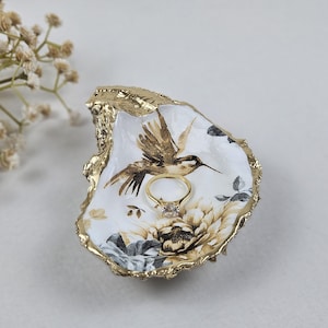 May include: A decorative oyster shell ring dish with a gold rim. The dish features a hummingbird and floral design in shades of brown, white, and gray. A gold ring with a square diamond sits inside the dish.