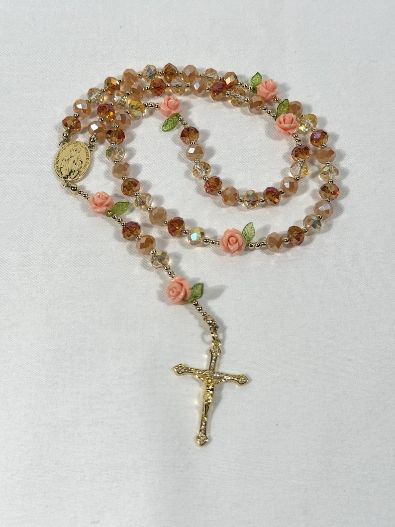 May include: A rosary featuring amber-toned beads and delicate pink rose embellishments. It includes a gold-coloured crucifix and a medallion with a religious figure. The beads are strung on gold-coloured wire.