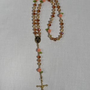 May include: A rosary featuring gold-coloured beads and a gold-tone crucifix. The beads are a mix of clear and amber hues, with small pink roses and green leaves. The rosary includes a gold-tone centrepiece with a religious image.