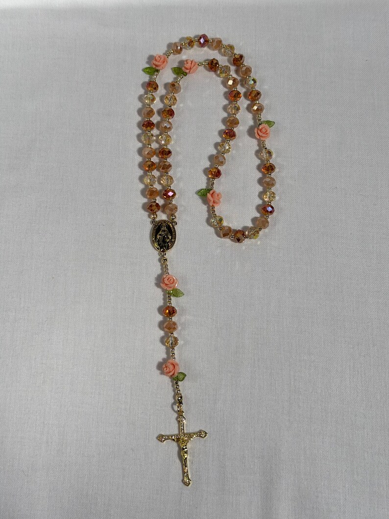 May include: A rosary featuring gold-coloured beads and a gold-tone crucifix. The beads are a mix of clear and amber hues, with small pink roses and green leaves. The rosary includes a gold-tone centrepiece with a religious image.