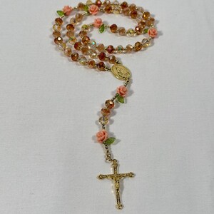 May include: A rosary featuring amber-coloured beads and gold-tone metal detailing. Small pink roses and green leaves are interspersed throughout. The rosary includes a gold crucifix and a medallion.
