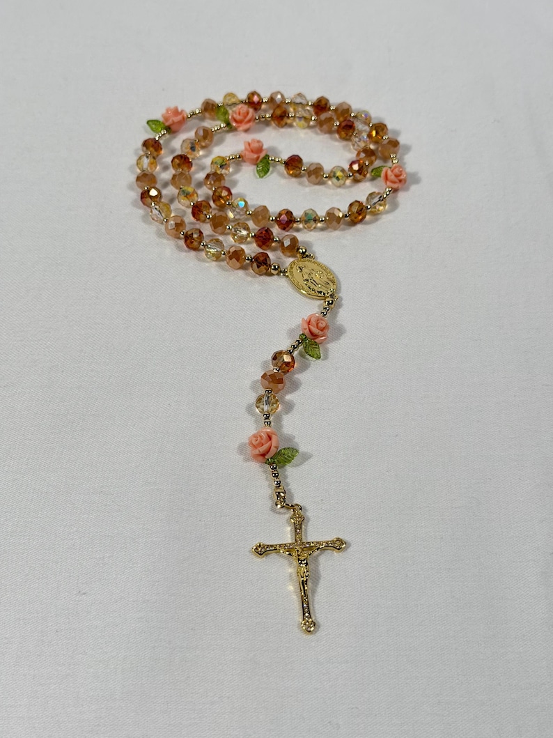 May include: A rosary featuring amber-coloured beads and gold-tone metal detailing. Small pink roses and green leaves are interspersed throughout. The rosary includes a gold crucifix and a medallion.