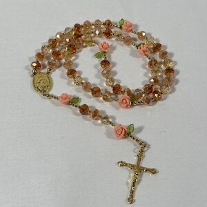 May include: A rosary featuring amber-coloured faceted beads and gold-tone metal details. It includes pink rose accents and a gold crucifix. The rosary also has a gold medal with an image.
