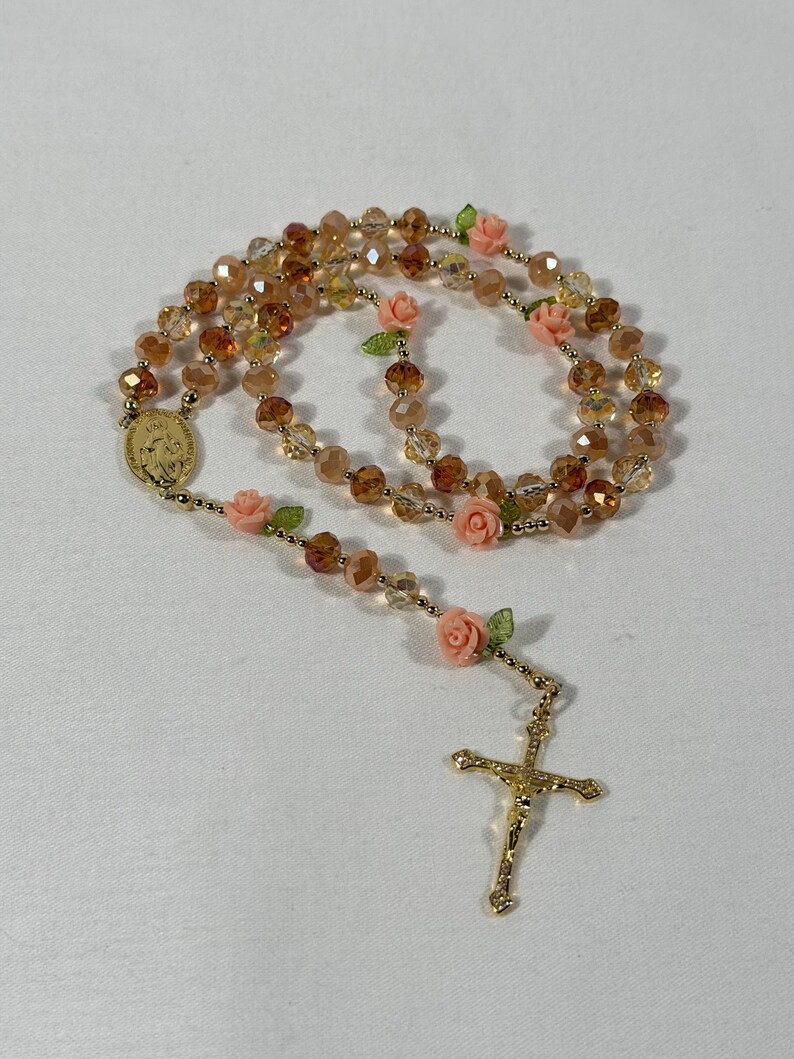 May include: A rosary featuring amber-coloured faceted beads and gold-tone metal details. It includes pink rose accents and a gold crucifix. The rosary also has a gold medal with an image.