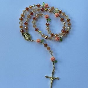 May include: A rosary featuring gold, amber, and brown faceted beads. Small peach-coloured rose accents are placed along the chain. A gold crucifix and a religious medallion are at the end of the chain.